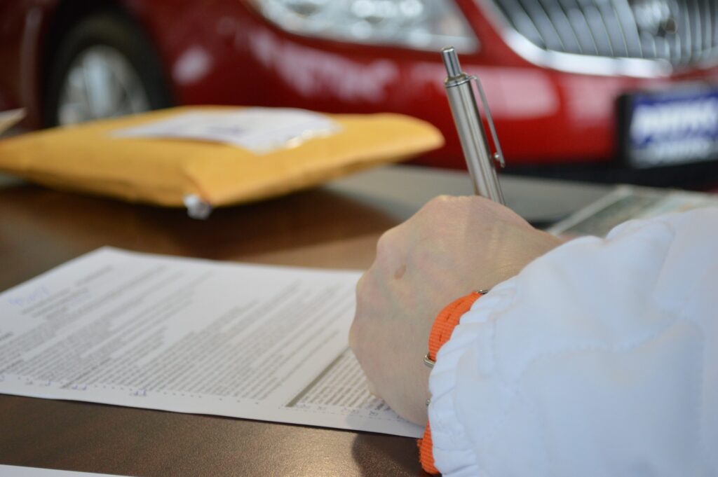 A hand signing some paperwork with a car in the background